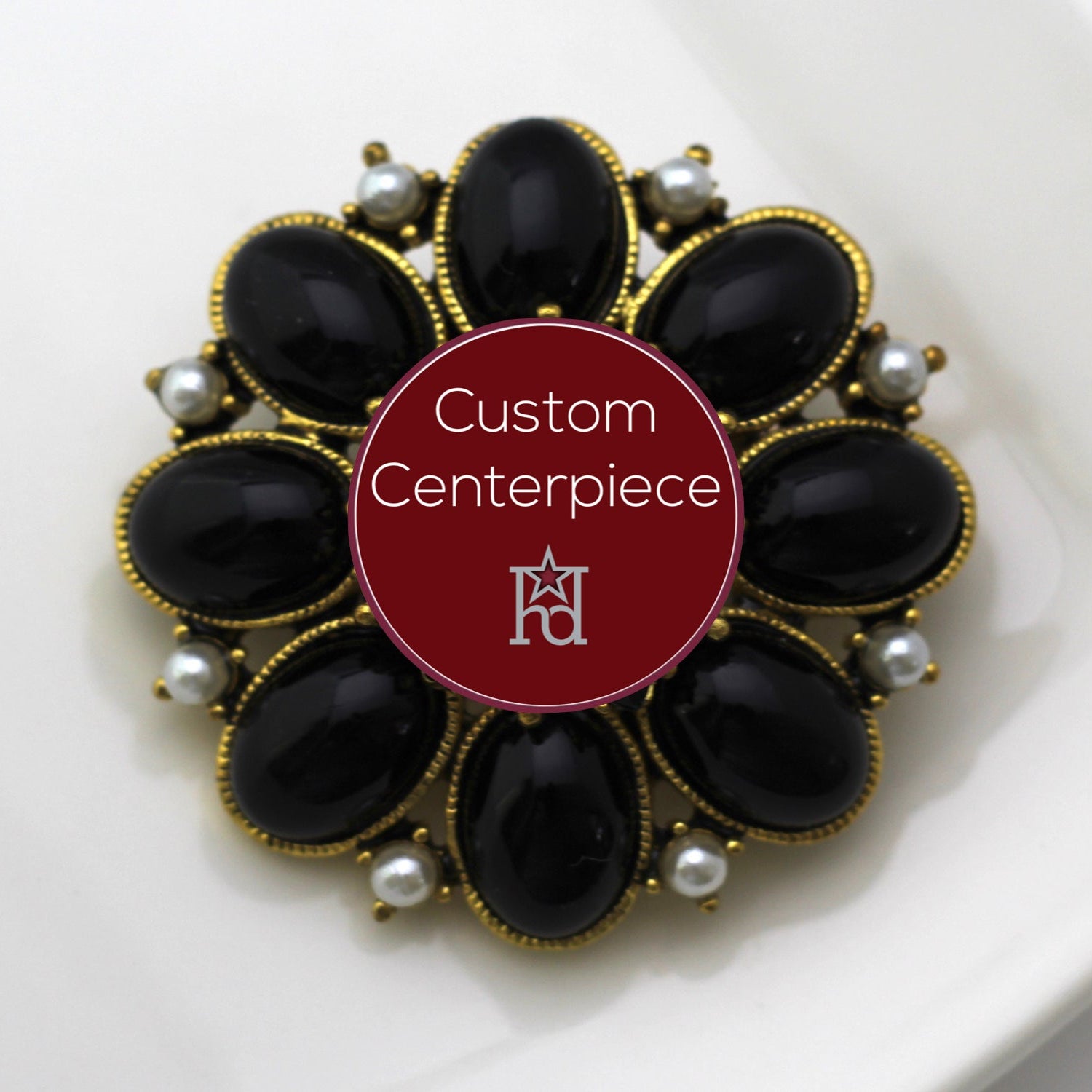 A gold brooch with black cabochons and faux pearls arranged in a floral pattern around a central red label with custom centerpiece text.