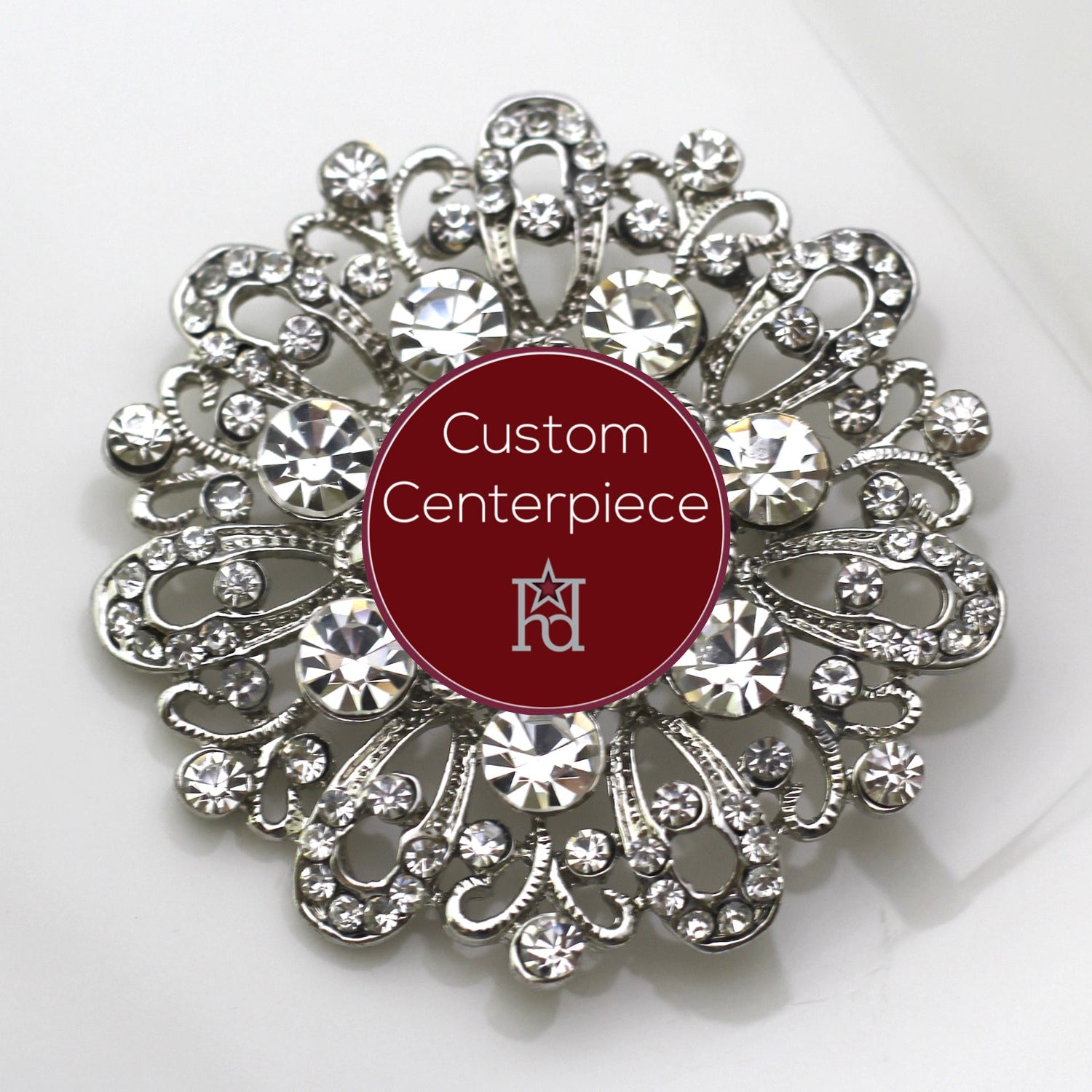 A silver brooch with clear crystals designed in a floral pattern, featuring a custom center piece with the text 'Custom Centerpiece' and the Hope Design logo.