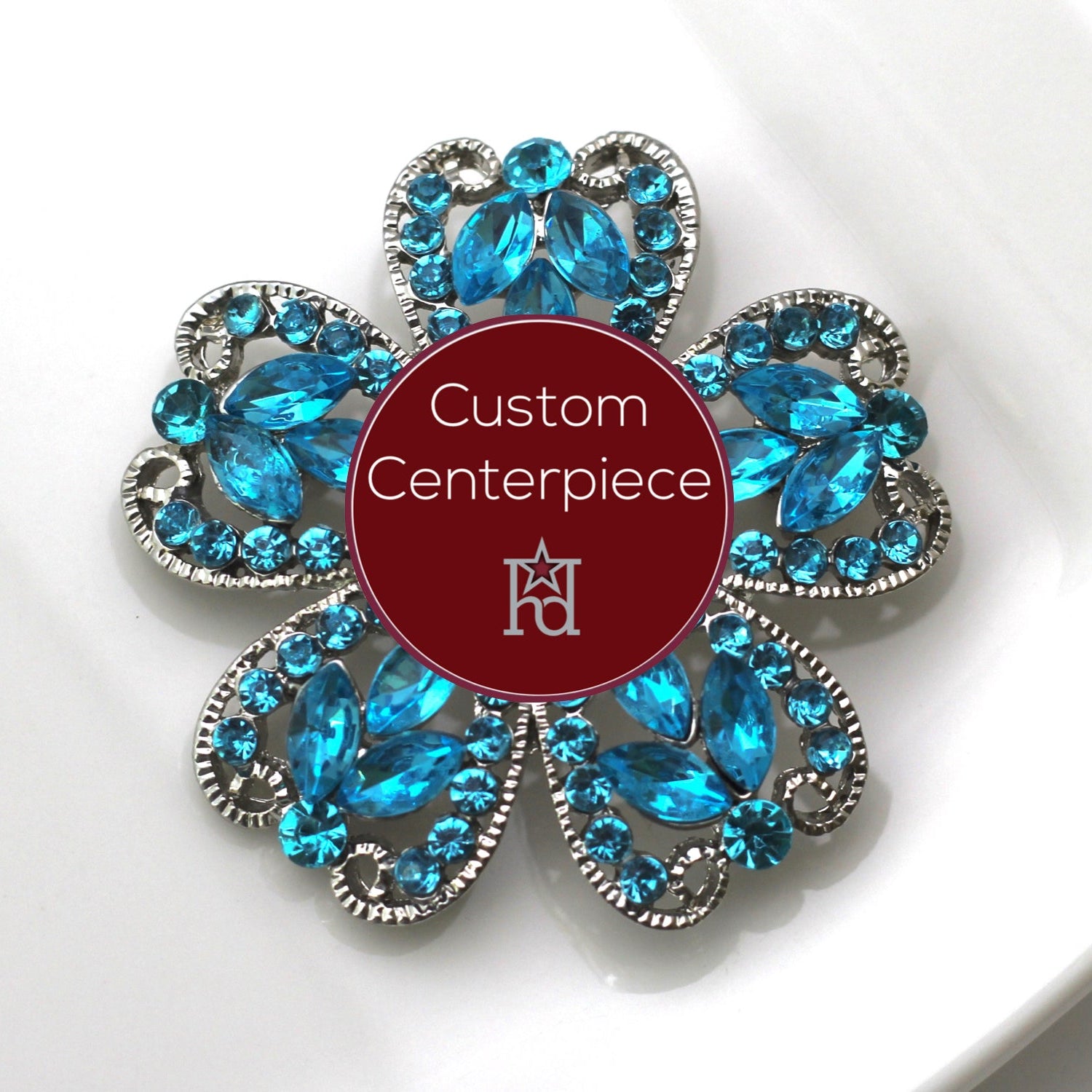 A silver brooch with light blue crystals designed in a floral pattern, featuring a central red label with the text 'Custom Centerpiece' on a white background.