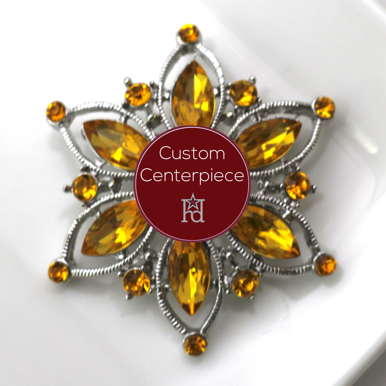 A custom-designed brooch with a silver base and citrine crystals, featuring a flower-like shape with a central larger citrine crystal surrounded by smaller ones, on a white background with a red 'H' logo in the corner.