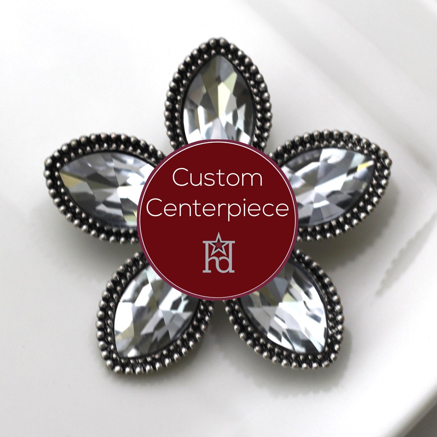 A silver brooch with clear crystals arranged in a flower pattern, displayed on a white surface with a custom centerpiece label.