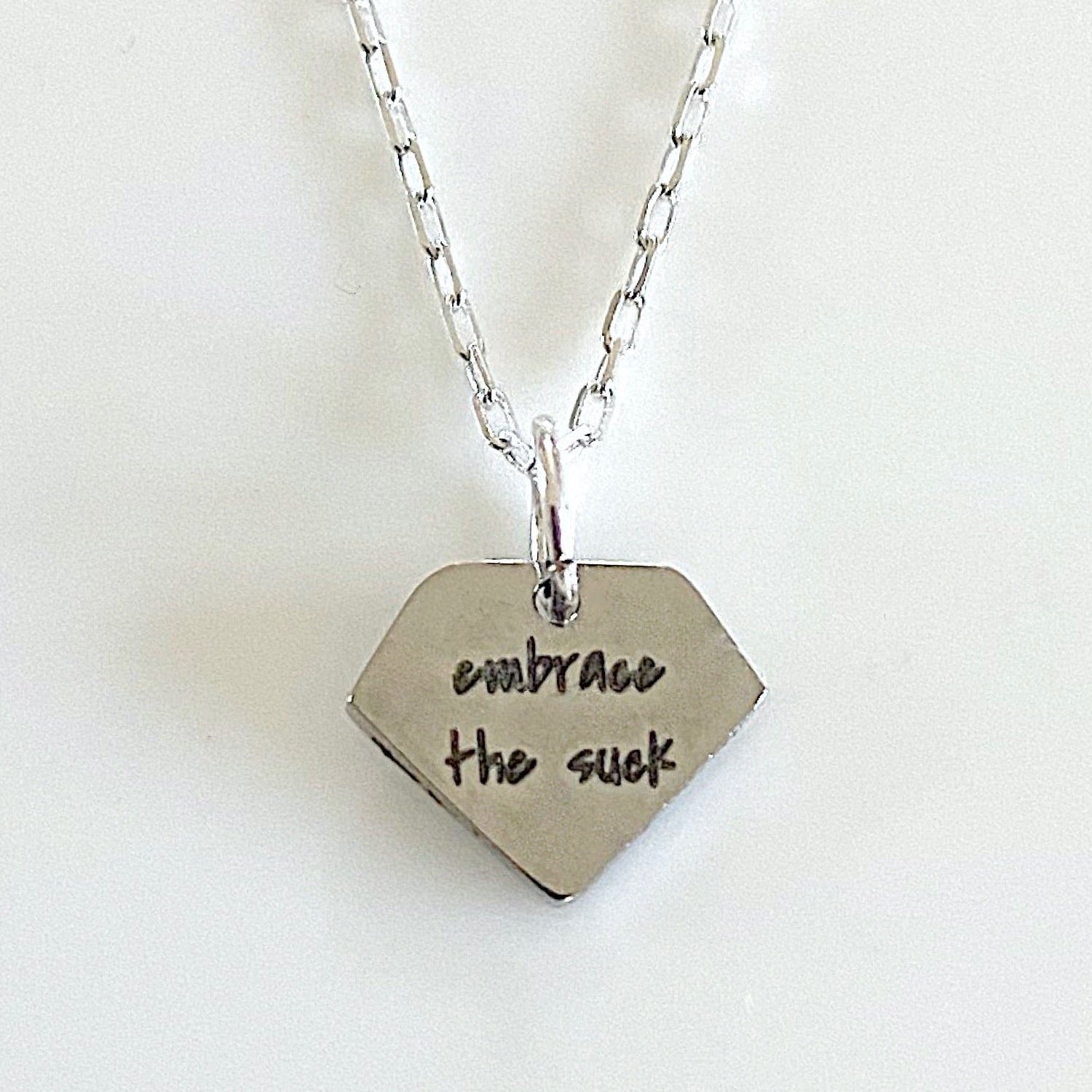 A silver-tone diamond pendant on a sterling silver chain with the phrase 'embrace the suck' engraved on it.