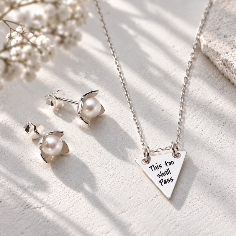 Silver necklace and matching stud earrings with pearl-like flower centers, styled on a light textured surface with soft shadows and small white flowers. The necklace pendant is a triangle engraved with the phrase 'This too shall pass'.