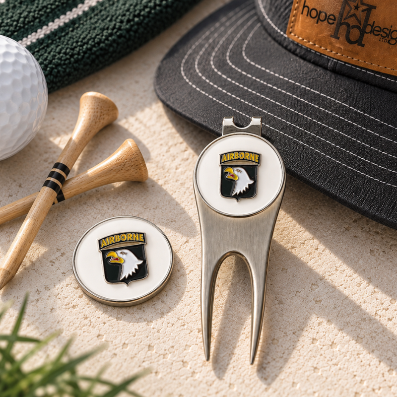 A silver metal golf divot tool and matching round golf ball marker set with an Arizona Wildcats eagle logo, styled on a sandy surface with a golf ball, wooden club heads, and a cap nearby.