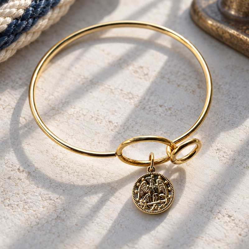 A gold-plated bangle bracelet with a small circular Saint Benedict medallion charm, displayed on a textured white surface with soft shadows.