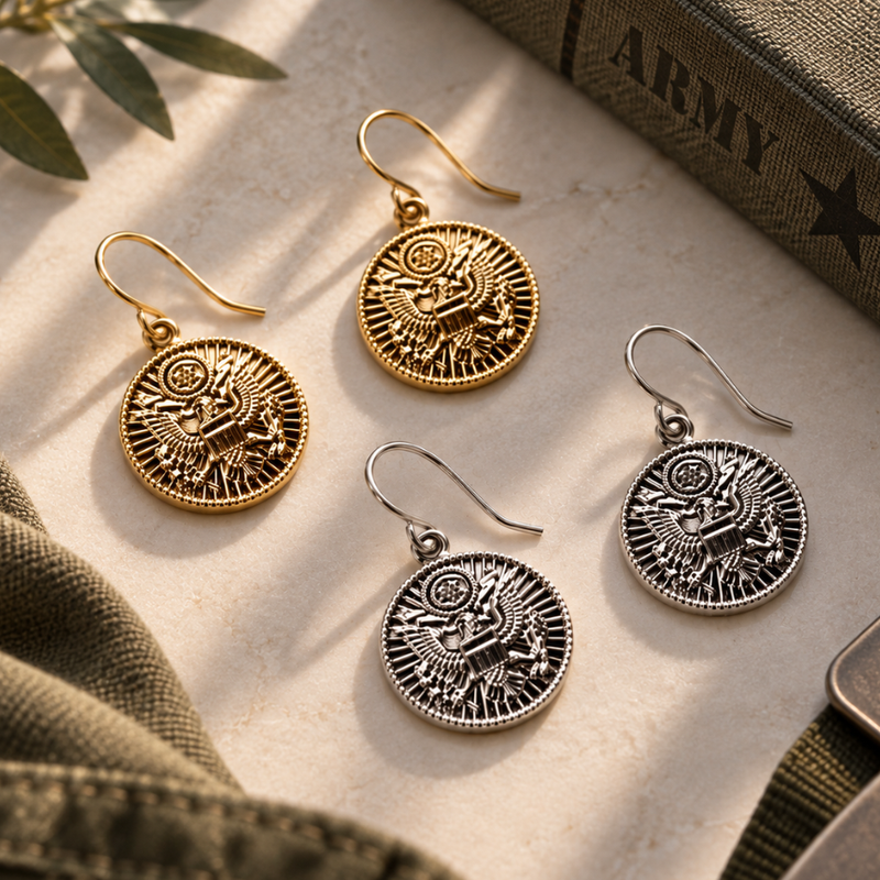 Four eagle-themed coin drop earrings in both gold and silver finishes, arranged on a textured beige surface with military-style fabric and a book in the background.