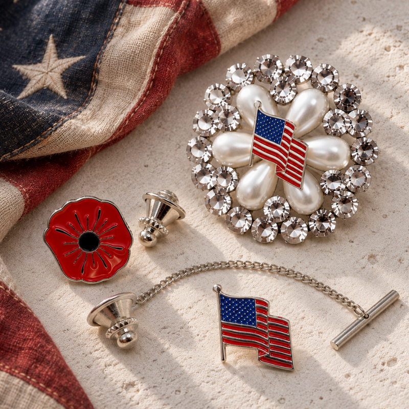 A styled flat-lay of patriotic American-themed accessories on a light surface with a vintage flag. The set includes a flower-shaped brooch with faux pearls and rhinestones surrounding an American flag, a red poppy pin, American flag tie tack.