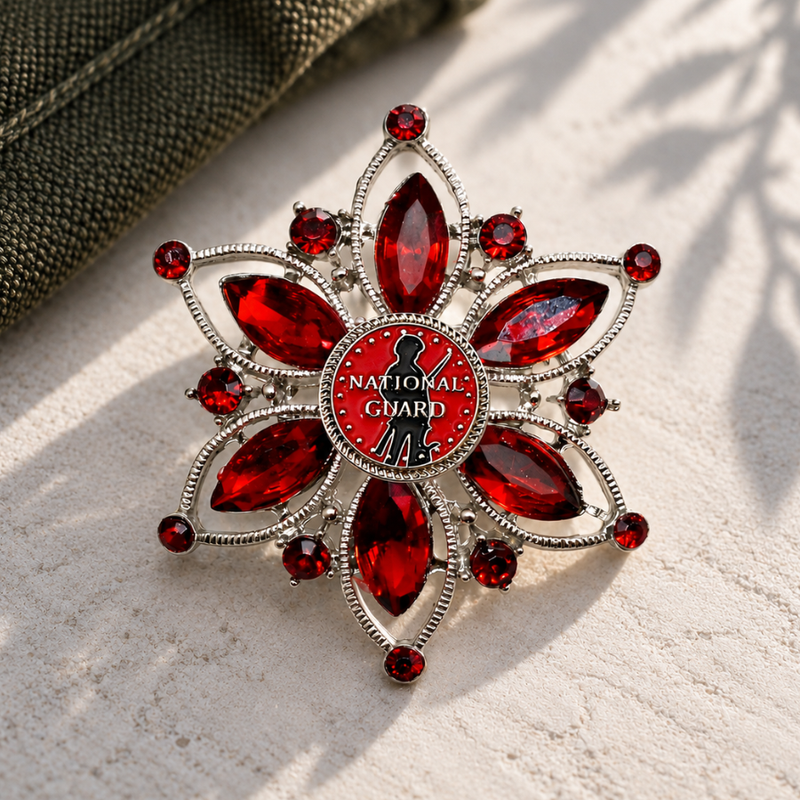 A silver-tone floral brooch with red faceted gemstones and a central red circle featuring the National Guard emblem and text, photographed on a light textured surface with a dark fabric corner in the upper left.