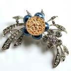 Decorative brooch with blue pearls, gold emblem, and silver leaves on a white background