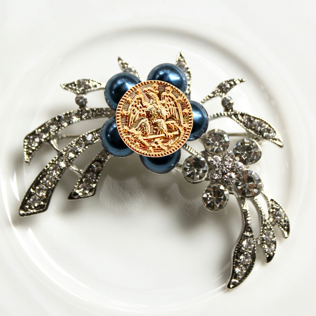 Decorative brooch with blue beads, gold emblem, and silver leaves on a white background