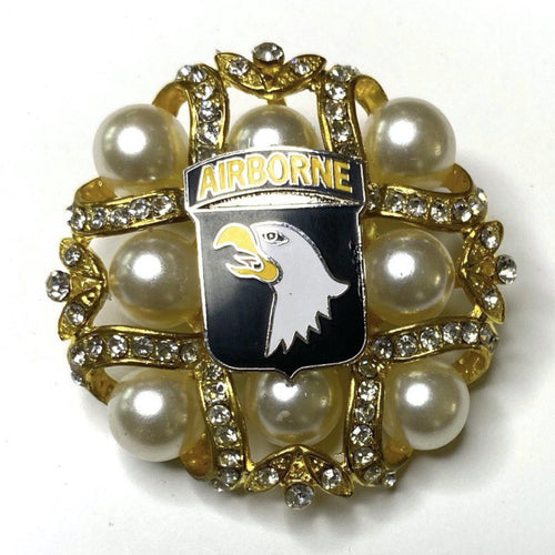 A gold-colored brooch featuring the 101st Airborne Division insignia with a bald eagle head, set against a background of pearl-like beads and rhinestones.