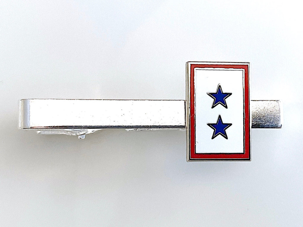 A tie clip with a blue star service flag design on a gold or silver metal clip with a red and white border.