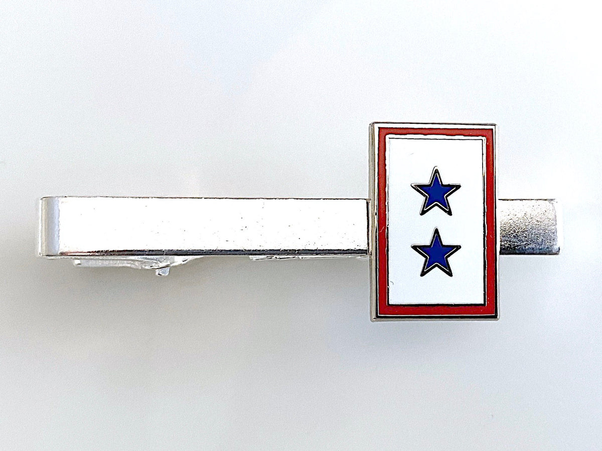 A tie clip with a blue star service flag design on a gold or silver metal clip with a red and white border.