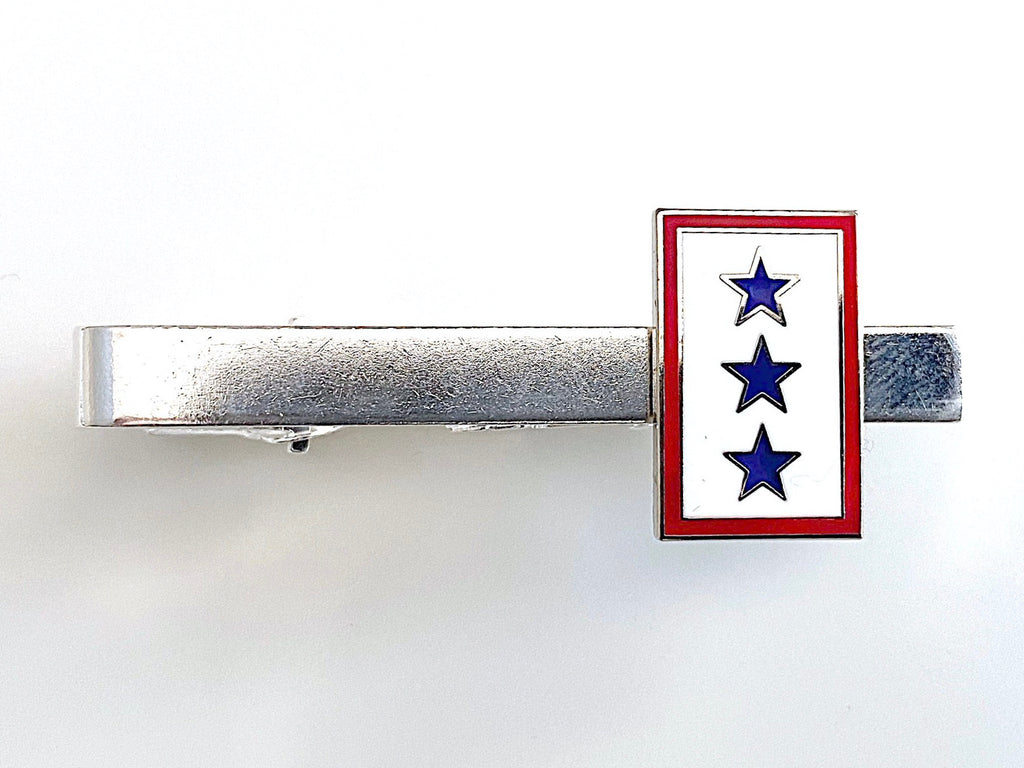 A tie clip with a blue star service flag design on a gold or silver metal clip with a red and white border.