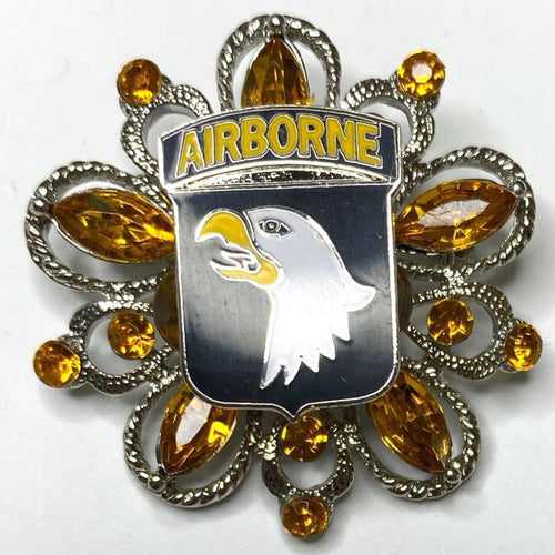A brooch featuring the 101st Airborne Division insignia with a bald eagle head, set against a background of amber-colored crystals.