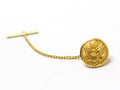 A gold-tone tie tack with a round button design featuring an emblem, attached to a small chain.