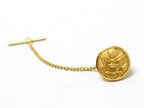A gold-tone tie tack with a round button design featuring an emblem, attached to a small chain.