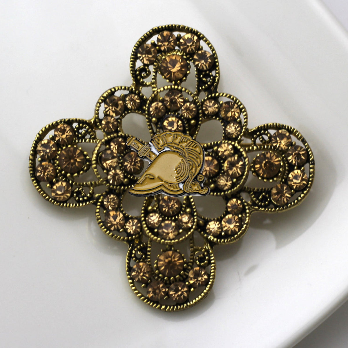 An ornate vintage-style brooch featuring a gold-tone central helmet motif surrounded by layered filigree circles set with brown faceted rhinestones, displayed on a white surface.