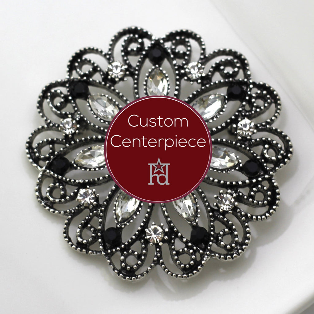 A custom-designed brooch with silver, black, and clear crystals, featuring a heraldic design, placed on a white surface with the brand name 'Hope Design Ltd.' visible.