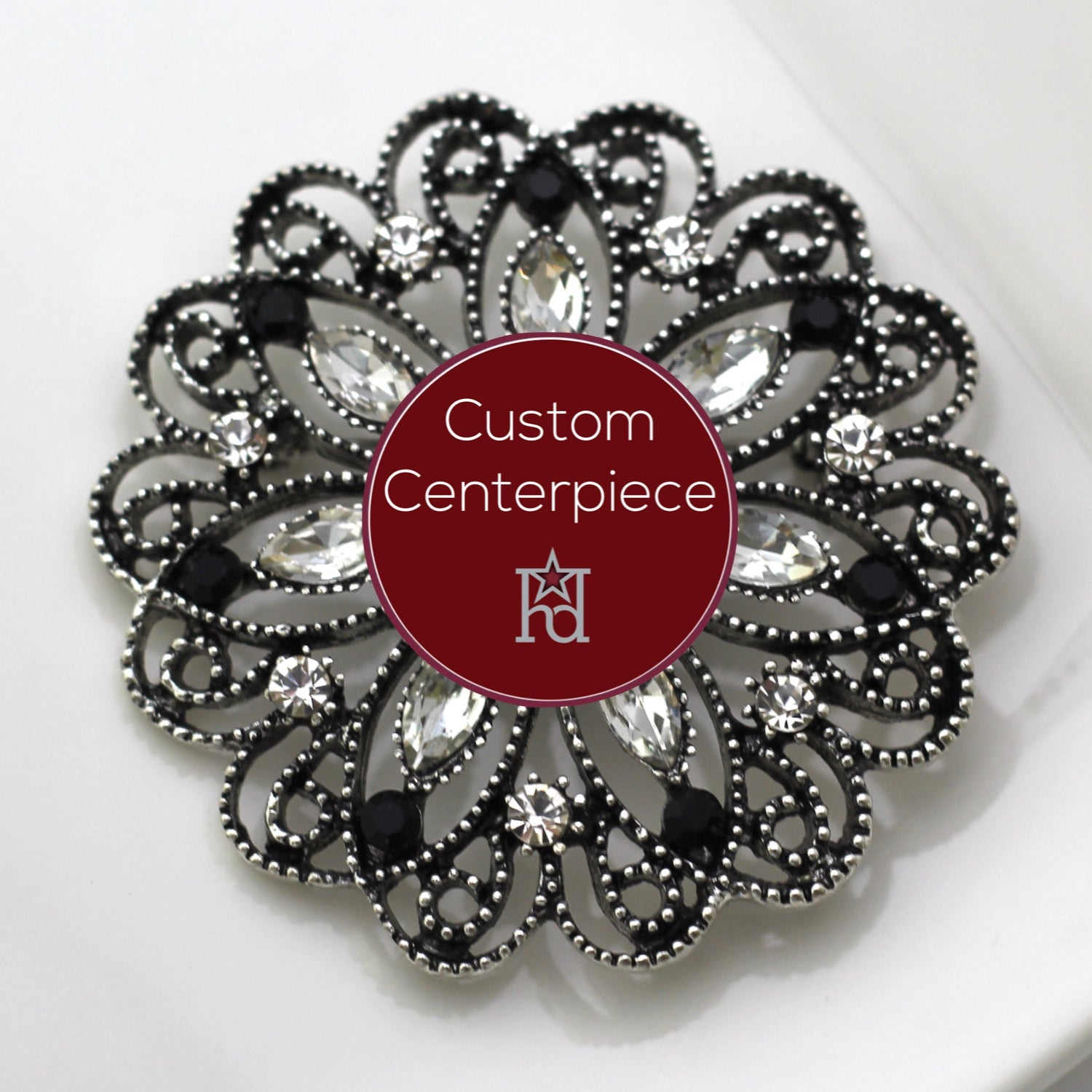 A custom-designed brooch with silver, black, and clear crystals, featuring a heraldic design, placed on a white surface with the brand name 'Hope Design Ltd.' visible.