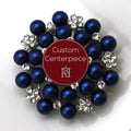 A round silver brooch with clear crystals and royal blue faux pearls arranged in a pattern around a custom centerpiece.