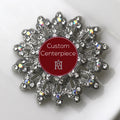 A silver brooch with clear crystals designed in a floral pattern with a custom centerpiece that has a red background and a white emblem.