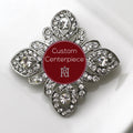 A silver brooch adorned with clear crystals and featuring a custom red centerpiece with a white letter 'h'.