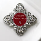 A silver brooch adorned with clear crystals and featuring a custom red centerpiece with a white letter 'h'.