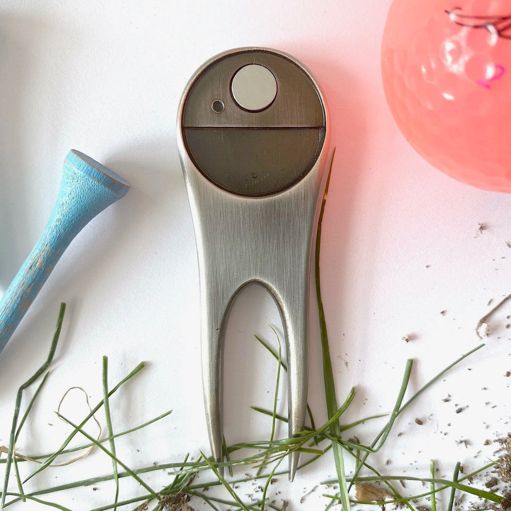 Metallic golf ball marker on a white surface with grass and a pink object in the background