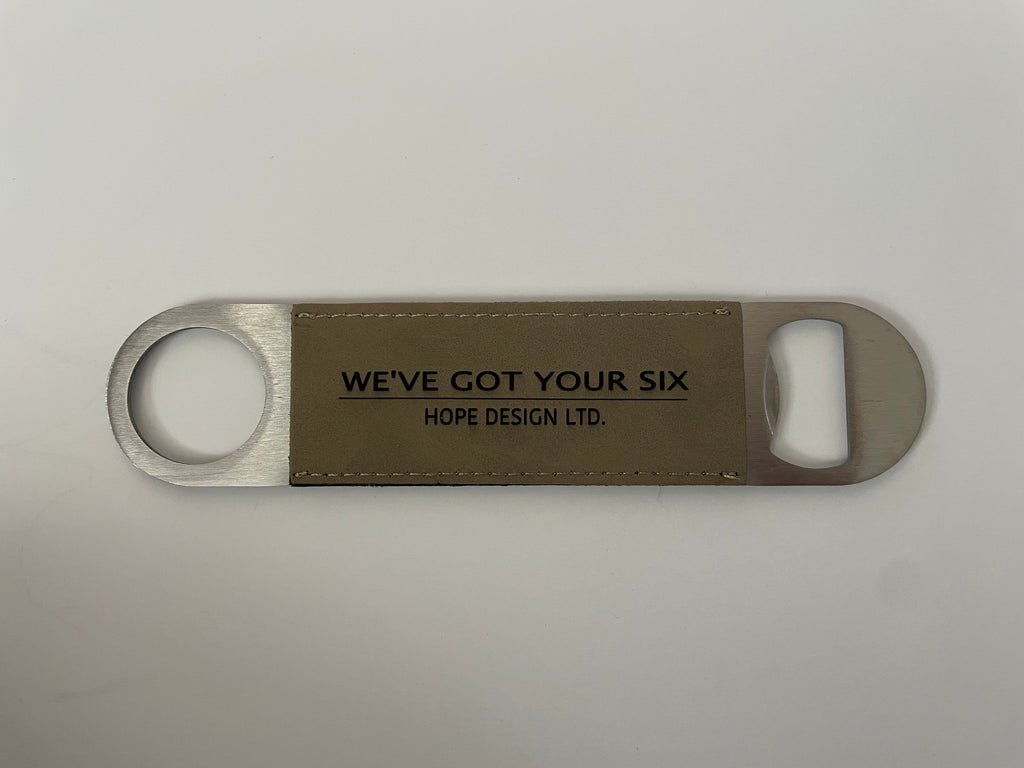 A bottle opener with a silver metal body and a brown faux leather strap around the middle. It has text on the strap that reads 'WE'VE GOT YOUR SIX'.