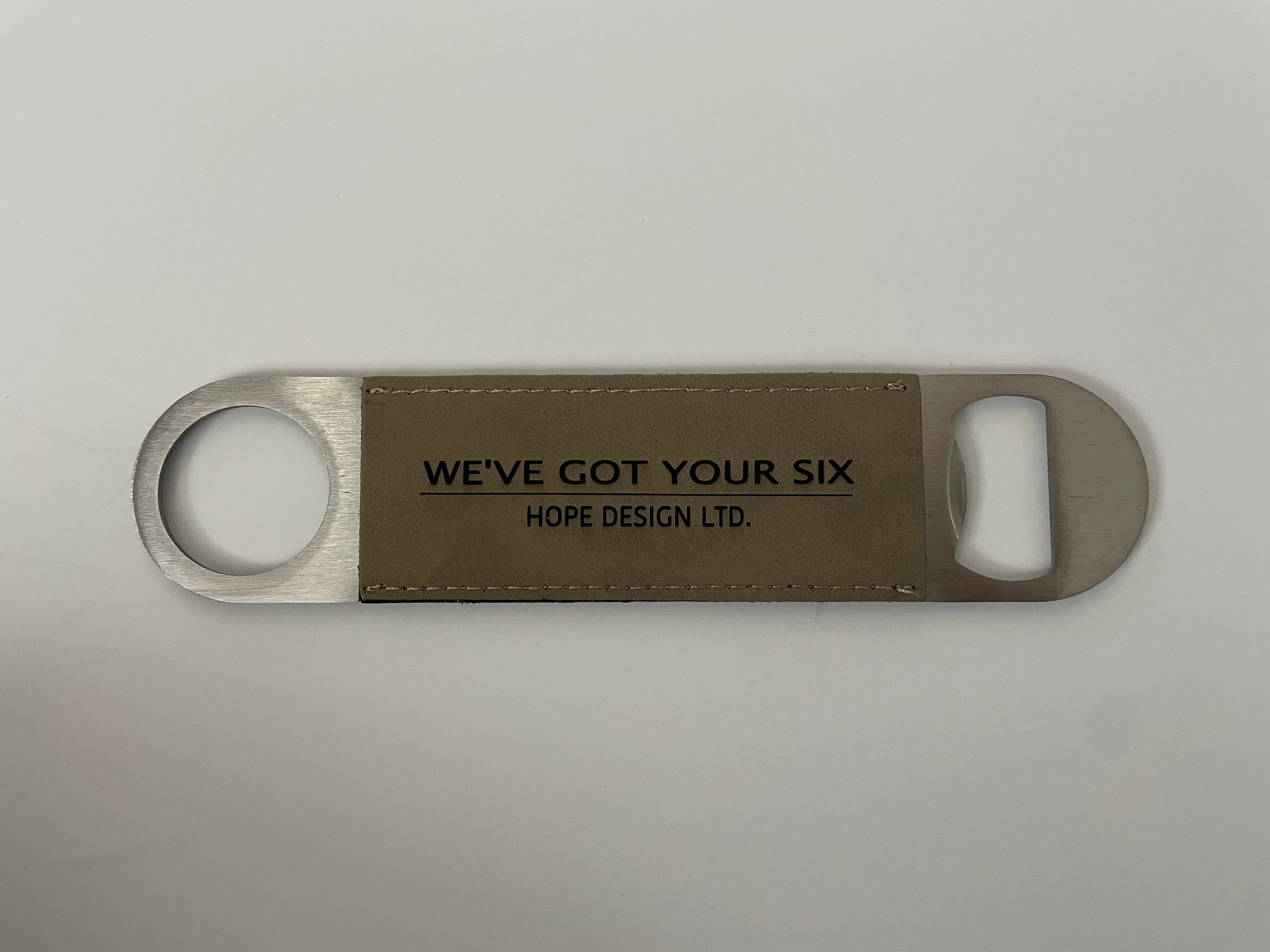 A bottle opener with a silver metal body and a brown faux leather strap around the middle. It has text on the strap that reads 'WE'VE GOT YOUR SIX'.