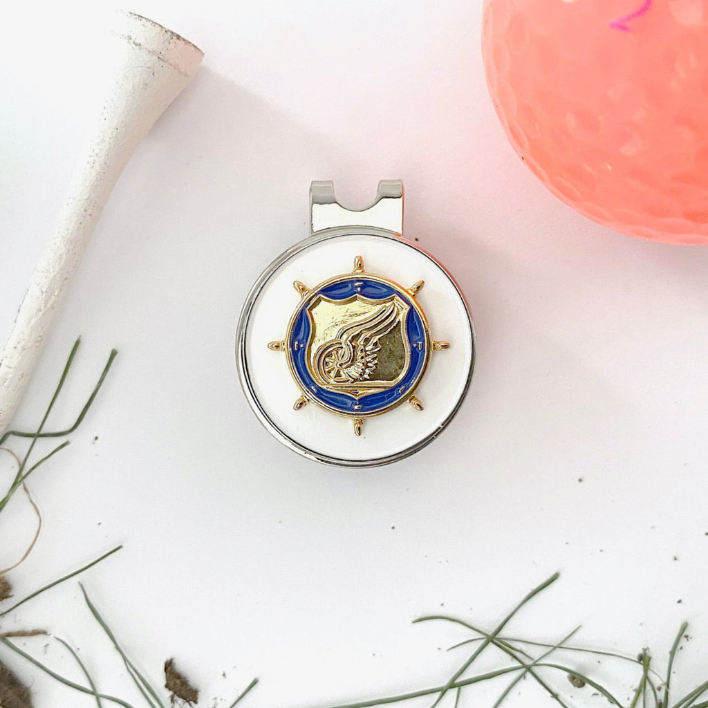Golf ball marker with emblem on a white surface with pink object and green leaves