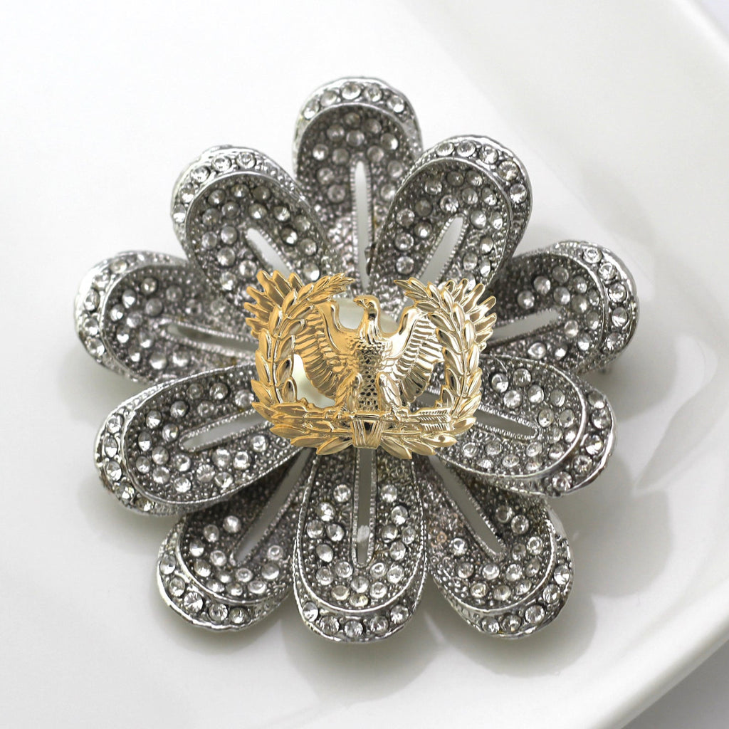 A silver-toned flower-shaped brooch with clear rhinestone detailing, photographed on a white surface.